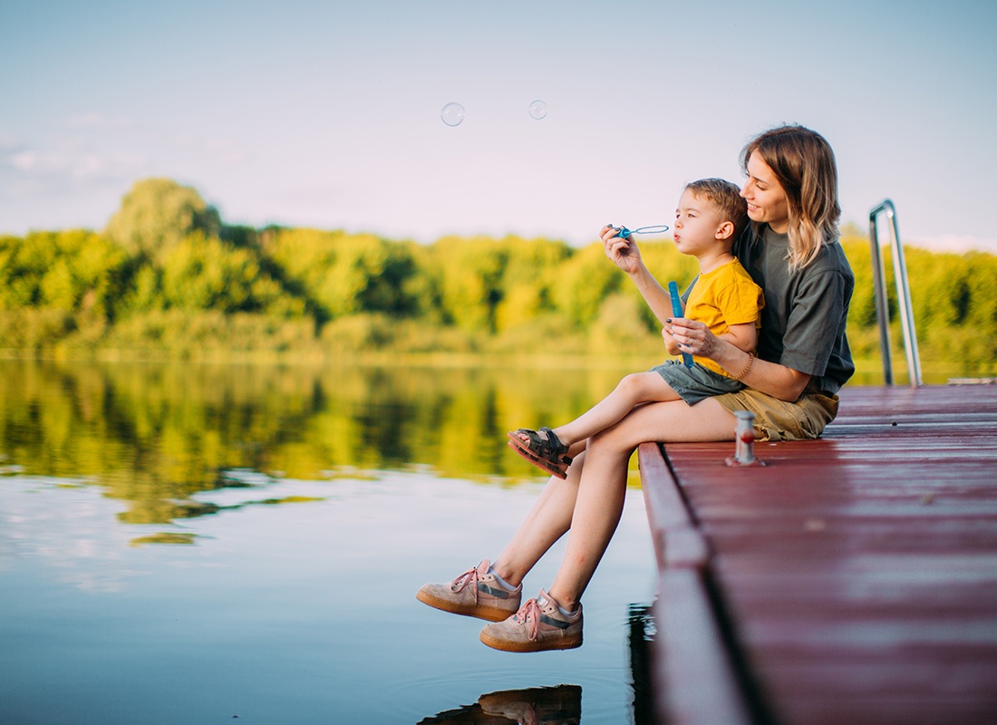 About Our Agency - Portrait of a Mother Sitting with her Son on the Edge of a Wooden Dock by the Lake While Blowing Bubbles