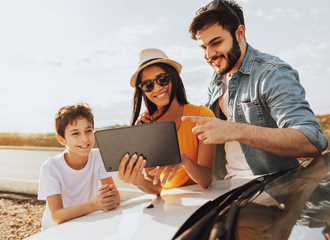 Insurance Solutions - Cheerful Family with a Younger Son Looking at a Tablet While Standing Outside Their Car Parked on the Side of the Road During a Summer Road Trip